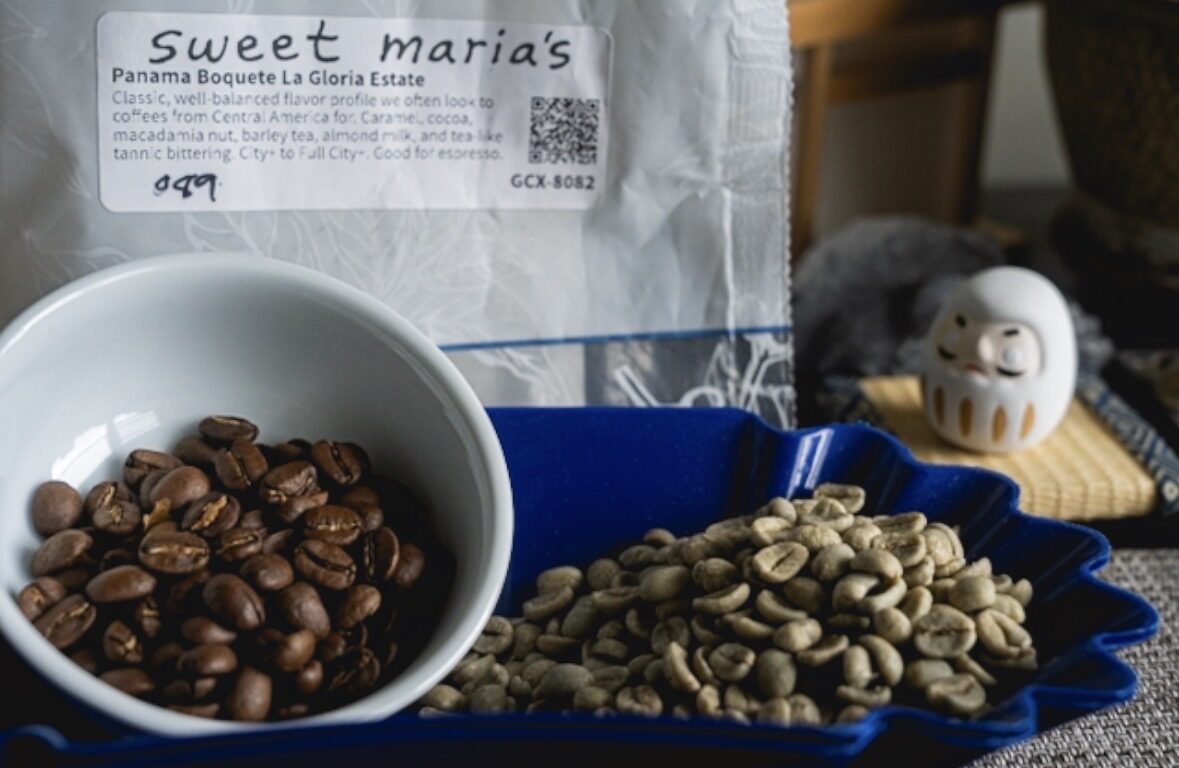 A white bowl of dark roasted beans next to a blue tray of pale green unroasted beans, with a Sweet Maria's coffee bag behind them.
