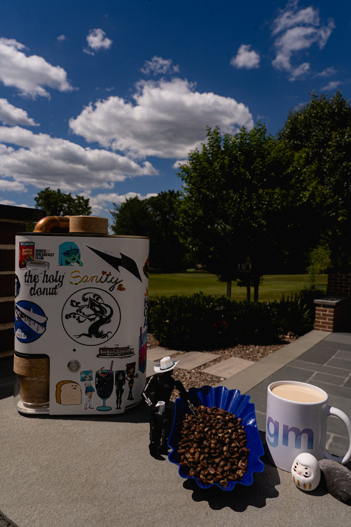 Ryan's home roasting setup outdoors: a sticker-covered roaster, a blue tray of fresh-roasted beans, a 'gm' mug, and a little robot in a cowboy hat.