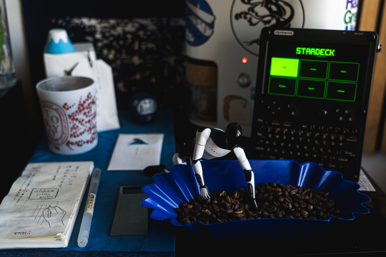 A small humanoid robot reclining on a blue tray of roasted beans next to a handheld device with a glowing green 'STARDECK' screen.